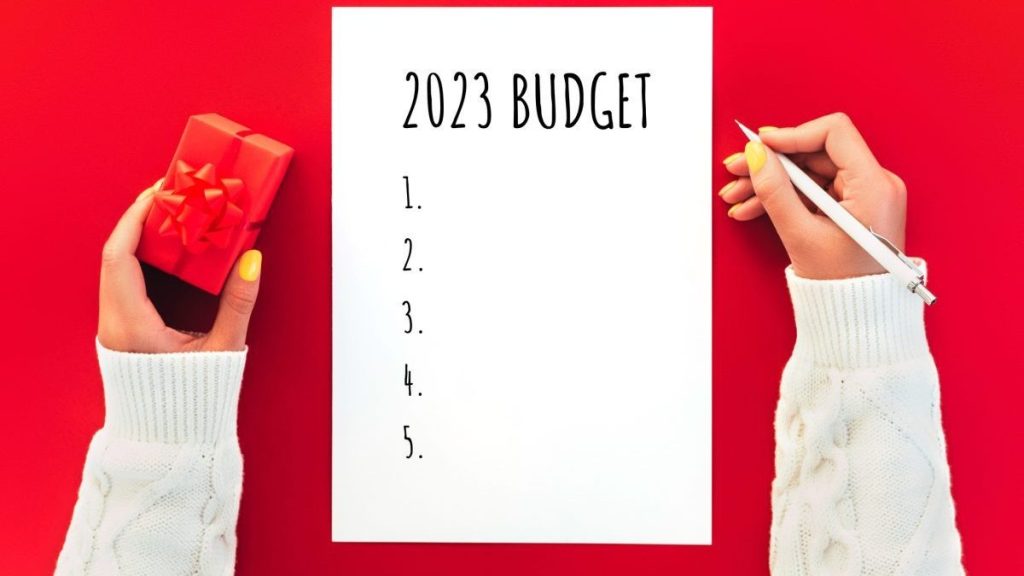 Woman's hands holding little red gift and writing budget for 2023 year on white paper.