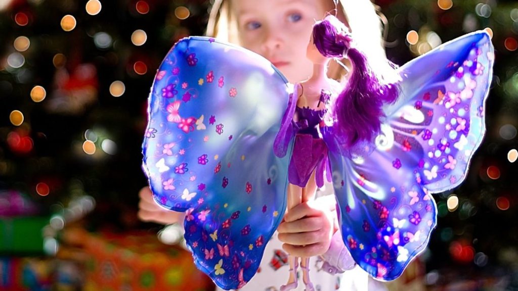 A three year old girl on Christmas morning in front of a Christmas tree with her new fairy doll. Background is filled with colorful Christmas tree lights.