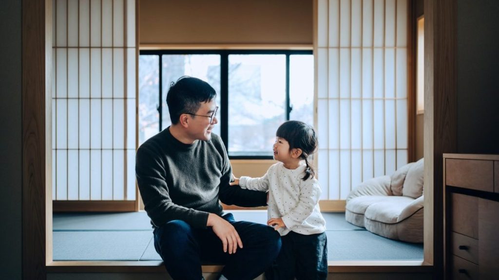 Young Asian father with his cute little daughter sitting on tatami mat in a traditional Japanese style bed and breakfast, chatting and enjoying father and daughter bonding time.