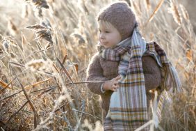 Beautiful toddler boy, dressed in knitted outfit with hat and flannel scarf, playing in frosty morning forest