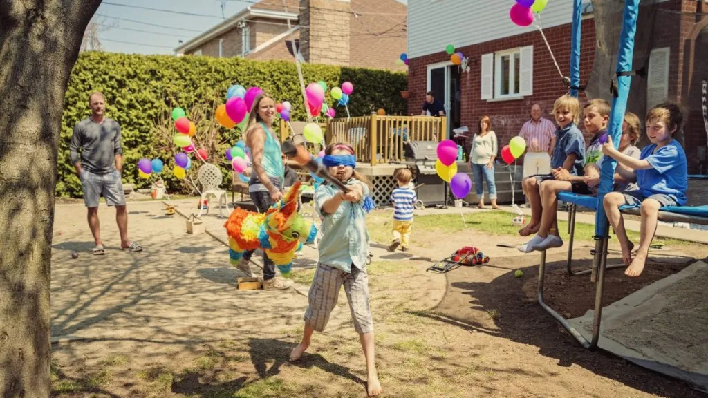 Little girl hitting pinata at birthday party in backyard. Lots of colorful ballons in the background with family and guests. Some kids are sitting on the trampoline watching with interest.