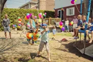 Little girl hitting pinata at birthday party in backyard. Lots of colorful ballons in the background with family and guests. Some kids are sitting on the trampoline watching with interest.