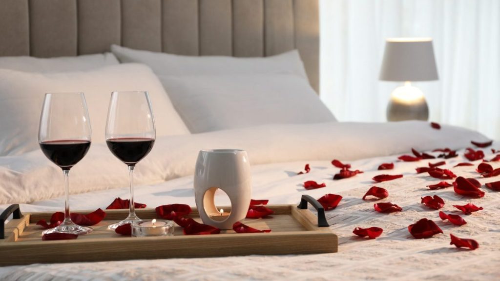 Tray with glasses of red wine, candles and rose petals on bed in room