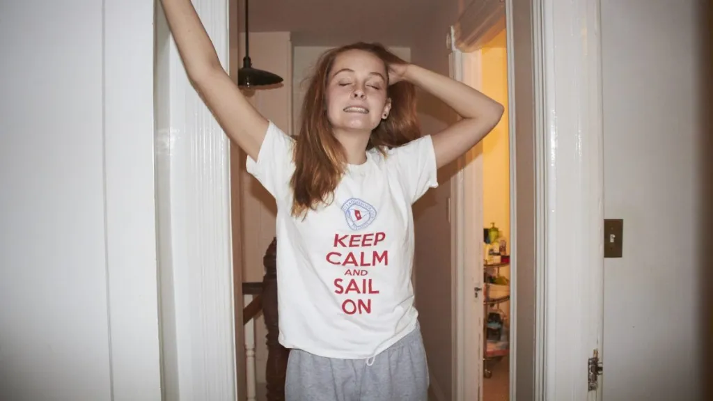 Shot of a teenage girl standing in a hallway of a home. She has long red hair. She has one arm up on the wall and the other hand is in her hair. She has her head thrown back and her eyes are closed and she is smiling. She is wearing a t shirt with a funny saying on it. She looks like she is sleepy. She seems like she could be just waking up or going to sleep.