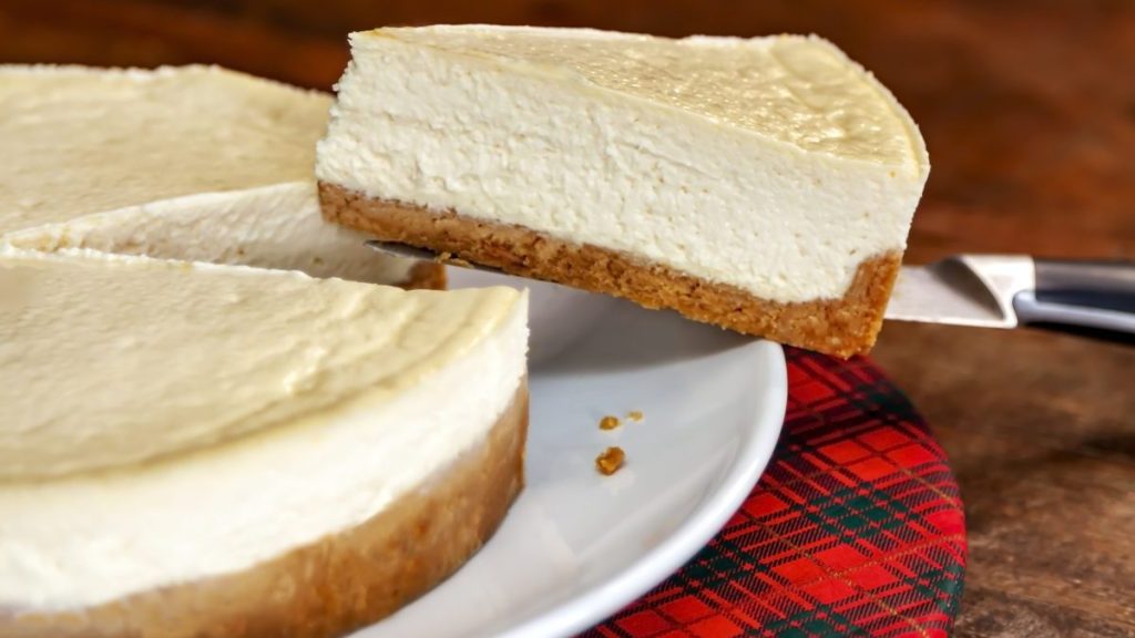 Removing a slice of the cheesecake with the help of a knife, the dessert is on a plate and on a plaid sideboard, all on the rustic wooden table. This dessert is not typical of Brazil, but is very appreciated by the Brazilian people.