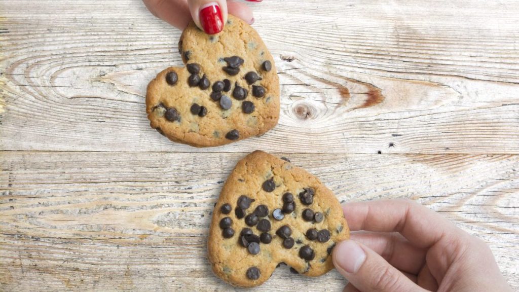 Young couple holding chocolate cookies in shape of hearts against wooden table, as love symbol