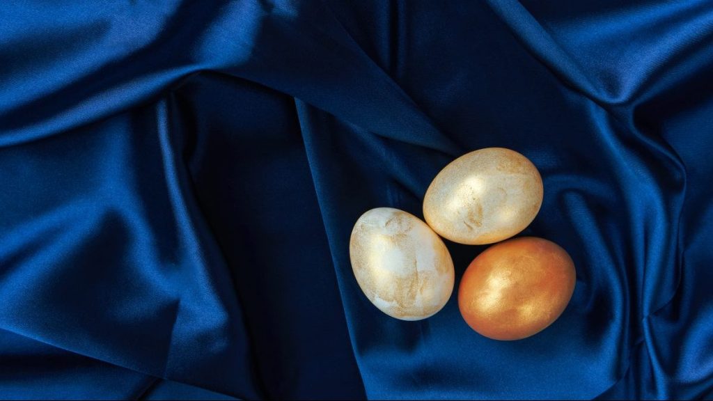 Three white and gold colored Easter eggs on blue satin background with copy space. Spring coming, eco-friendly products concept.