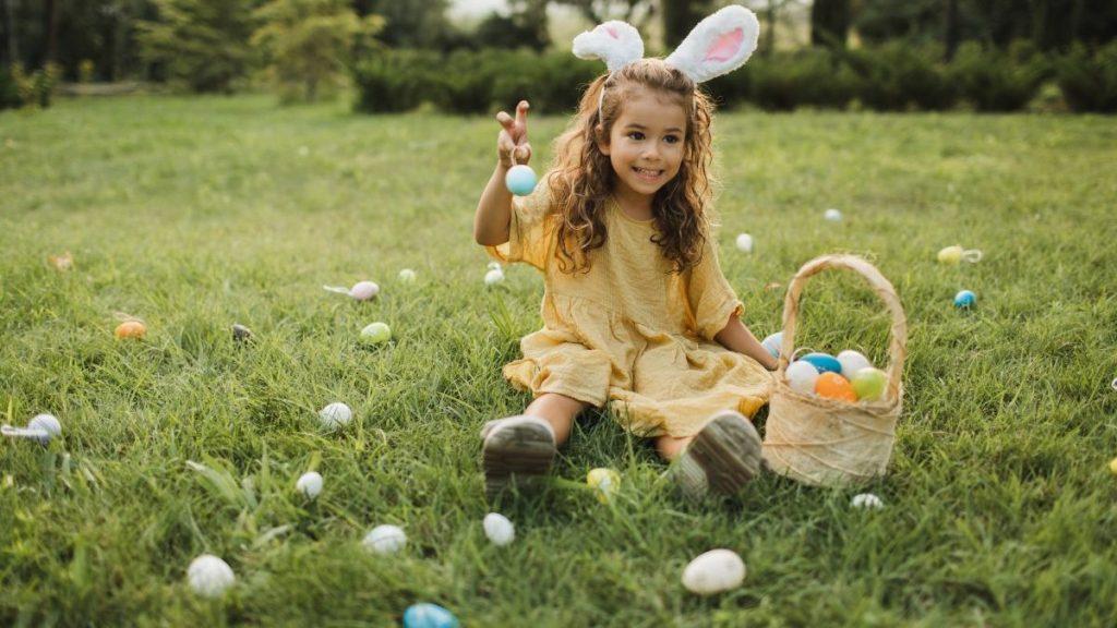 Cute little girl with bunny ears holding an easter egg basket and finding eggs in the grass on green lawn. Little girl wearing fluffy Bunny ears gathering colorful egg in park in wicker basket. Easter concept.