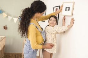 Young pregnant woman and her little daughter hanging pictures on the wall of the baby's bedroom
