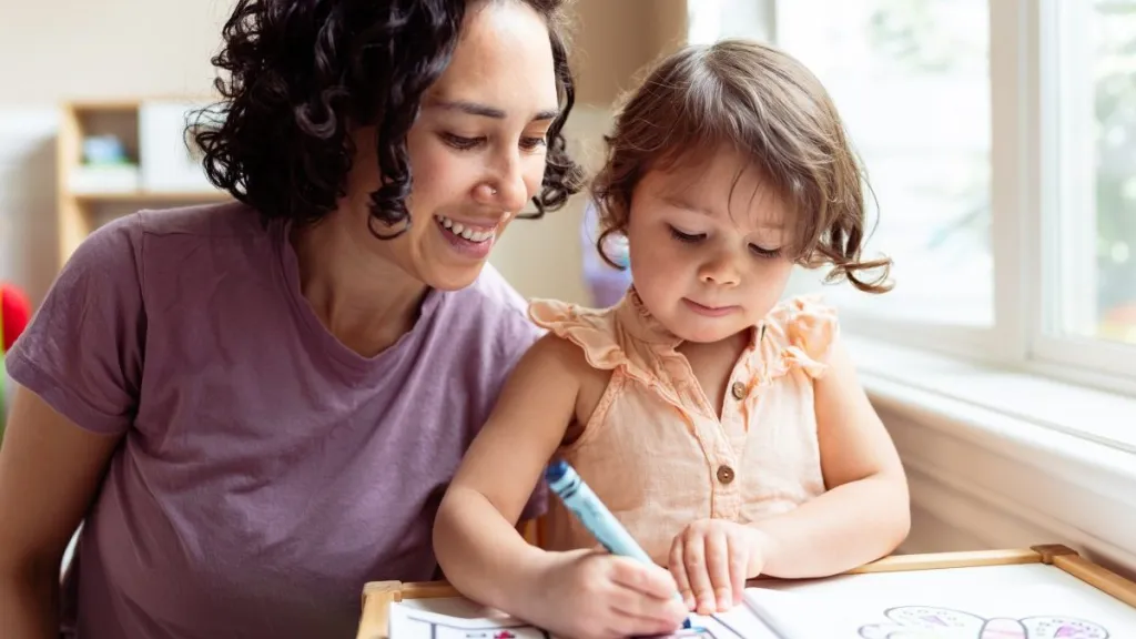 An Eurasian woman who is applying Montessori education techniques while homeschooling her preschool age daughter smiles encouragingly as the young girl colors with a crayon.