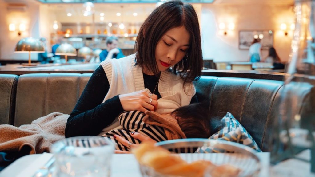 Beautiful young mother breastfeeding her baby in public while having lunch in restaurant.