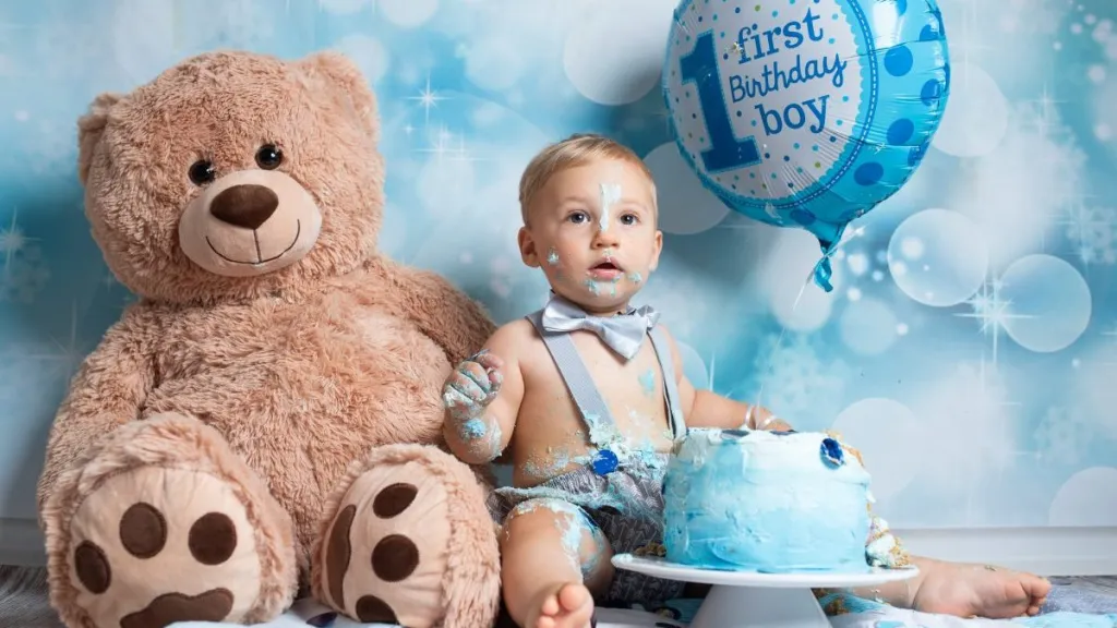 First Birthday: Young toddler posing for messy cake smash photoshoot with teddy bear and balloon in front of blue decorated background.