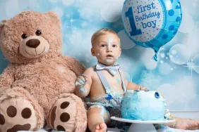 First Birthday: Young toddler posing for messy cake smash photoshoot with teddy bear and balloon in front of blue decorated background.