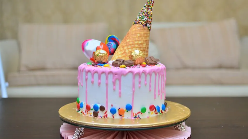 A cute pink birthday cake with an ice cream cone waffle / wafer on top with sprinkles chocolate and lollipop on top for a girl's birthday
