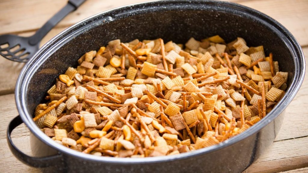 This is a shot of a party mix containing cereal, peanuts and pretzels in a roaster pan on an old wooden table.