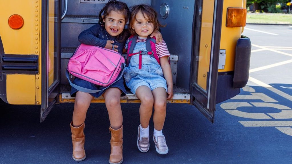 An Hispanic girl with down syndrome and her best girl friend who is Eurasian embrace while sitting on the steps of a school bus and smile with excitement before spending the day at school together.