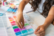 A girl sits at her kitchen table making bracelets for a fundraiser, carefully stringing beads and tying knots with a smile on her face, hoping to make a positive impact.